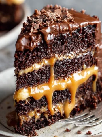 Homemade slice of chocolate caramel cake with moist chocolate layers, gooey caramel filling, glossy ganache topping, and chopped nuts, served on a rustic plate with warm lighting.