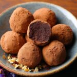 A bowl of cocoa-dusted homemade chocolate truffles, with one bitten to reveal a rich, smooth center, next to a cup of coffee on a dark cloth.