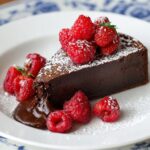 Homemade slice of flourless chocolate cake topped with fresh raspberries and dusted with powdered sugar, served on a white plate with a blue floral rim and a small pool of melted chocolate at the base.