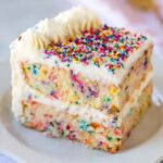 Homemade slice of Funfetti cake with vanilla frosting and colorful sprinkles on a white plate.