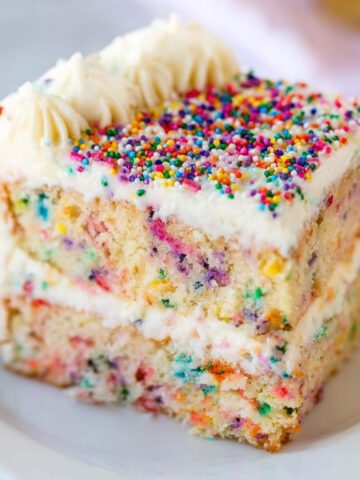Homemade slice of Funfetti cake with vanilla frosting and colorful sprinkles on a white plate.