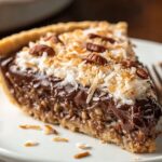 A homemade slice of German chocolate pie topped with toasted coconut and pecans, with a glossy chocolate filling and flaky golden crust on a white plate.