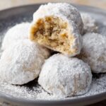 Homemade Italian wedding cookies on a plate, coated in powdered sugar, with one broken open to show a soft, nutty, crumbly inside.