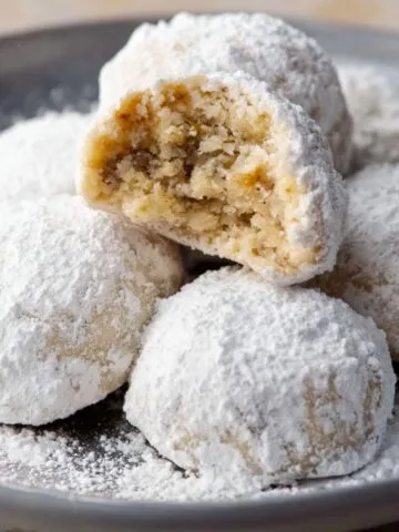 Homemade Italian wedding cookies on a plate, coated in powdered sugar, with one broken open to show a soft, nutty, crumbly inside.