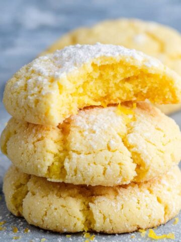 Stack of homemade lemon cookies on a gray surface, dusted with powdered sugar and lemon zest, with a bite taken from the top cookie revealing a soft, bright yellow center.