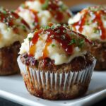 Homemade meatloaf cupcakes in white liners, topped with piped mashed potatoes, ketchup drizzle, and chopped chives on a white plate.