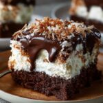 A homemade slice of Mounds Cake with moist chocolate cake, a thick coconut filling, rich chocolate ganache dripping down the sides, and toasted coconut sprinkled on top, served on a plate with a fork.