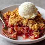 Homemade rhubarb dump cake with golden crumble topping and a scoop of vanilla ice cream, served in a white dish with a wooden spoon.