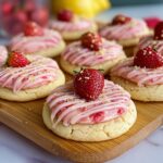 Homemade strawberry lemonade cookies with pink frosting, white drizzle, sprinkles, and fresh strawberry halves on top, arranged on a wooden board with lemons and strawberries in the background.