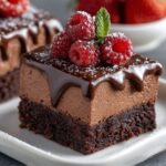 Homemade chocolate mousse brownies topped with glossy ganache, fresh raspberries, powdered sugar, and a mint leaf on a white plate.