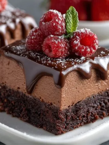 Homemade chocolate mousse brownies topped with glossy ganache, fresh raspberries, powdered sugar, and a mint leaf on a white plate.
