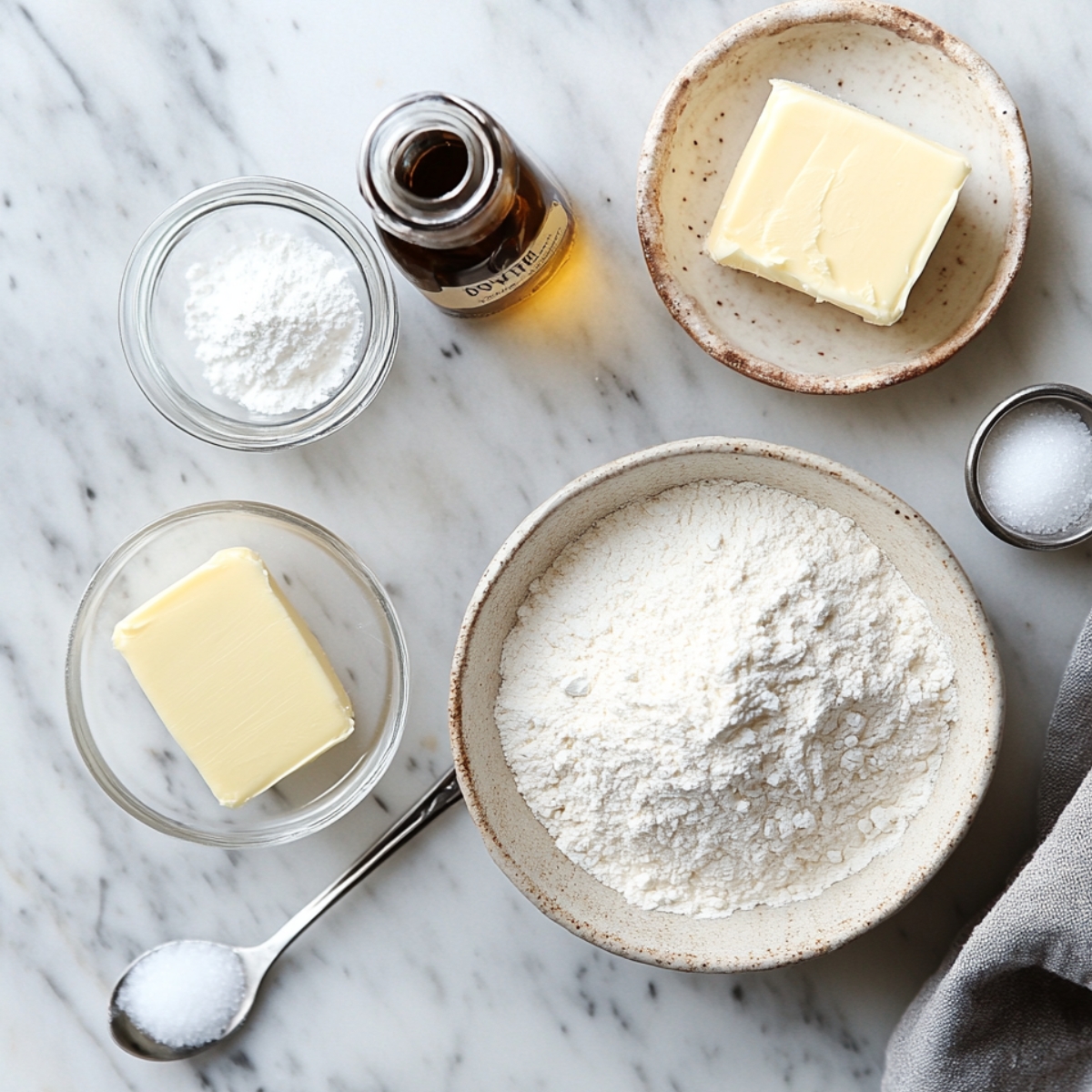 Flat lay of ingredients for edible sugar cookie dough on a marble surface: flour, butter, powdered sugar, vanilla extract, and salt.