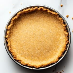 Golden baked graham cracker crust in a metal pie pan, sitting on a light marble surface with scattered crumbs around the edges, ready to be filled for a homemade s’mores pie.