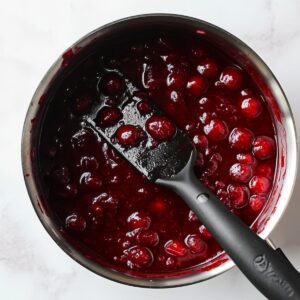 Thick cherry filling with whole cherries in a saucepan, stirred with a black spatula, part of homemade Black Forest brownie preparation.