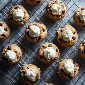 Freshly baked chocolate chip cookies cooling on a wire rack, each with a dollop of creamy cheesecake filling in the center. Cookies are golden brown with visible chocolate chips and a soft, thick texture.