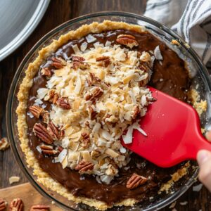 A homemade German chocolate pie in a glass dish with chocolate filling and a graham cracker crust, being topped with toasted coconut flakes and pecans using a red spatula.