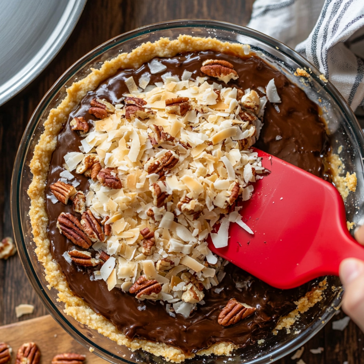 A homemade German chocolate pie in a glass dish with chocolate filling and a graham cracker crust, being topped with toasted coconut flakes and pecans using a red spatula.