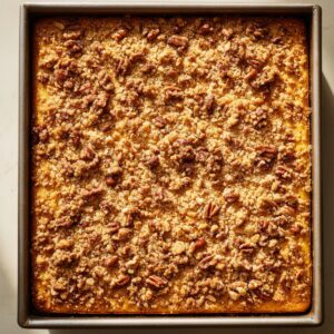 A realistic photo of a freshly baked pumpkin crunch cake in a square metal pan, with a golden-brown crumbly topping and toasted pecans scattered across the surface, lit by warm natural sunlight.