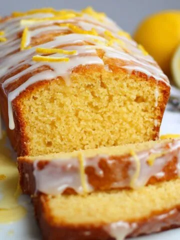 Best Lemon Drizzle Cake Recipe loaf on a wooden board, golden sponge sliced to show moist texture, topped with icing drizzle, lemon zest, and lemon slices, with blurred lemons in the background.