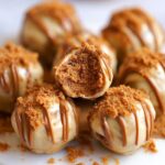 Homemade Biscoff truffles on a wooden board, coated in milk and white chocolate, drizzled with cookie butter, and topped with crushed Biscoff cookies. One is bitten to reveal a soft, golden center.