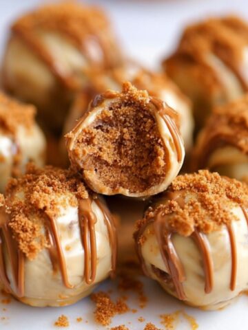 Homemade Biscoff truffles on a wooden board, coated in milk and white chocolate, drizzled with cookie butter, and topped with crushed Biscoff cookies. One is bitten to reveal a soft, golden center.