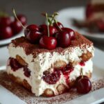 Close-up of homemade Cherry Amaretto Tiramisu with layers of mascarpone, soaked ladyfingers, and cherry filling, topped with cocoa powder and fresh cherries on a white plate.