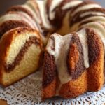 Cinnamon swirl bundt cake on a lace doily, with golden crumb, cinnamon sugar ridges, and white glaze drizzled over the top.