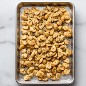 Golden homemade ranch oyster crackers on a baking sheet, sprinkled with herbs and black pepper.