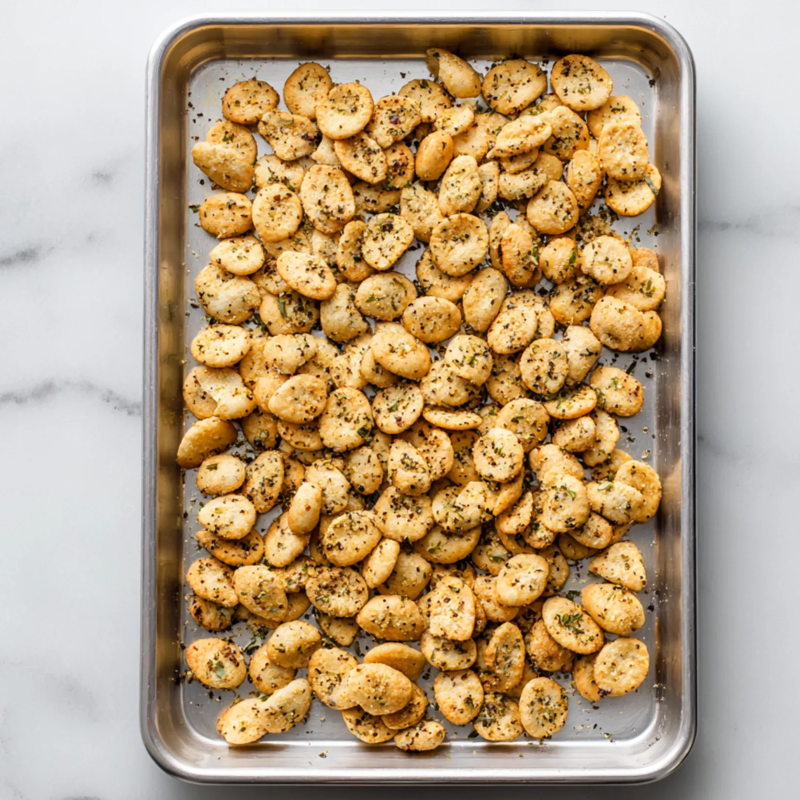 Golden homemade ranch oyster crackers on a baking sheet, sprinkled with herbs and black pepper.