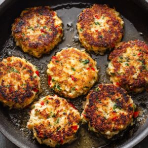 Golden brown homemade crab cakes frying in a black skillet, with crispy seared tops, chunks of crab meat, parsley, and red pepper visible.