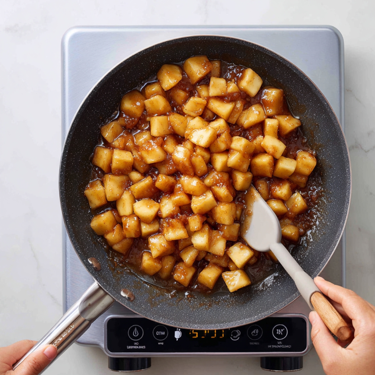 Diced apples in cinnamon-brown sugar sauce cooking in a skillet on a silver induction cooktop, stirred with a silicone spatula.
