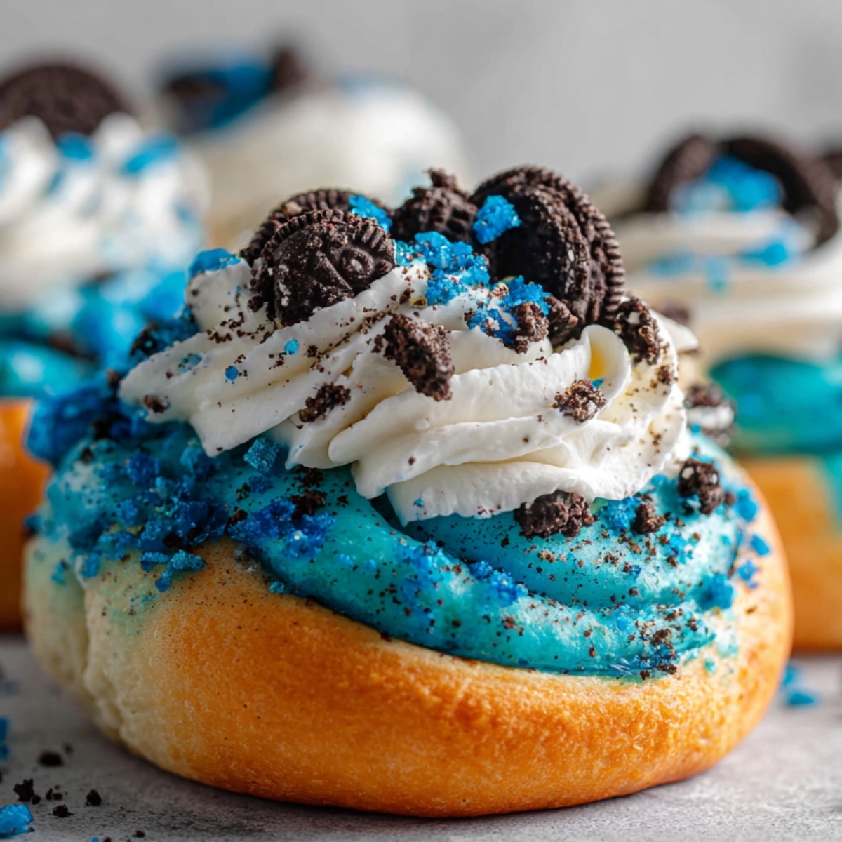 A soft, golden-brown Cookie Monster Cinnamon Rolls topped with bright blue frosting, a swirl of white whipped cream, crushed mini chocolate cookies, and blue sugar crystals. It’s homemade and detailed, with a fun Cookie Monster theme.