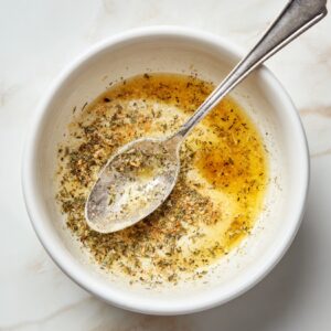 "White bowl with melted butter and ranch seasoning, stirred with a silver spoon, herbs visible in the mix.