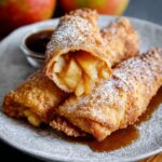 Easy Apple Pie Egg Rolls on a white plate, dusted with powdered sugar, filled with warm cinnamon-apple filling that oozes slightly.