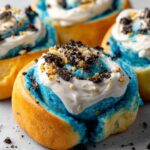 Homemade Cookie Monster cinnamon rolls with blue dough, white frosting, and crushed Oreos on top, sitting on a light surface.