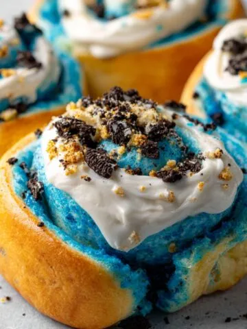 Homemade Cookie Monster cinnamon rolls with blue dough, white frosting, and crushed Oreos on top, sitting on a light surface.