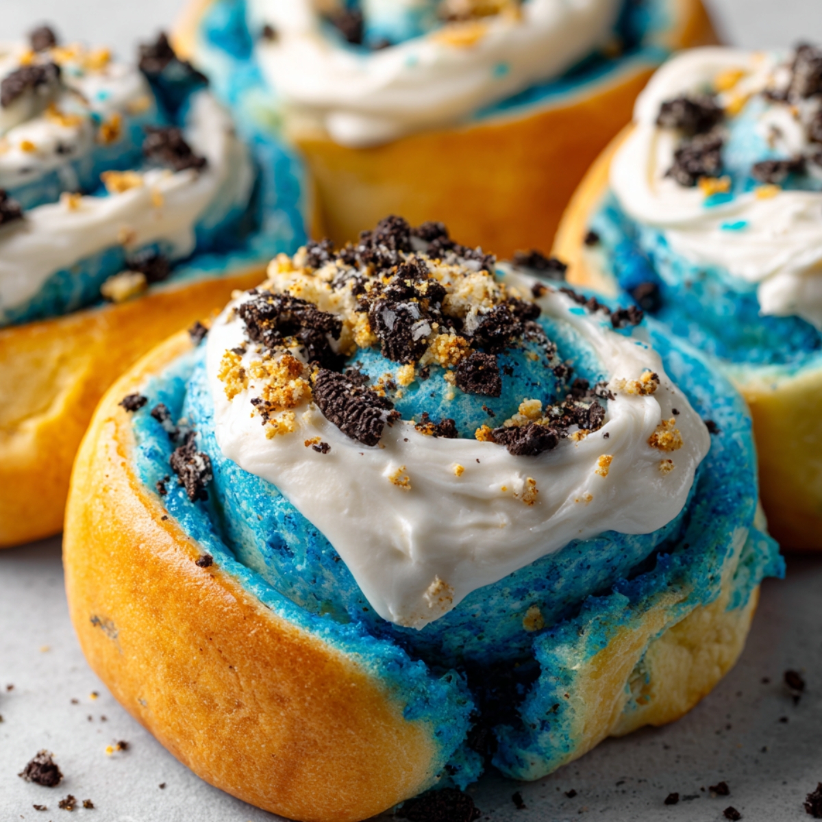 Homemade Cookie Monster cinnamon rolls with blue dough, white frosting, and crushed Oreos on top, sitting on a light surface.