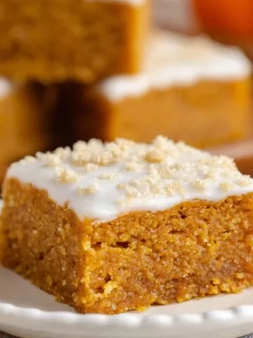 Moist homemade pumpkin bars topped with white frosting and sugar sprinkles, served on a scalloped ceramic plate with other bars in the background.