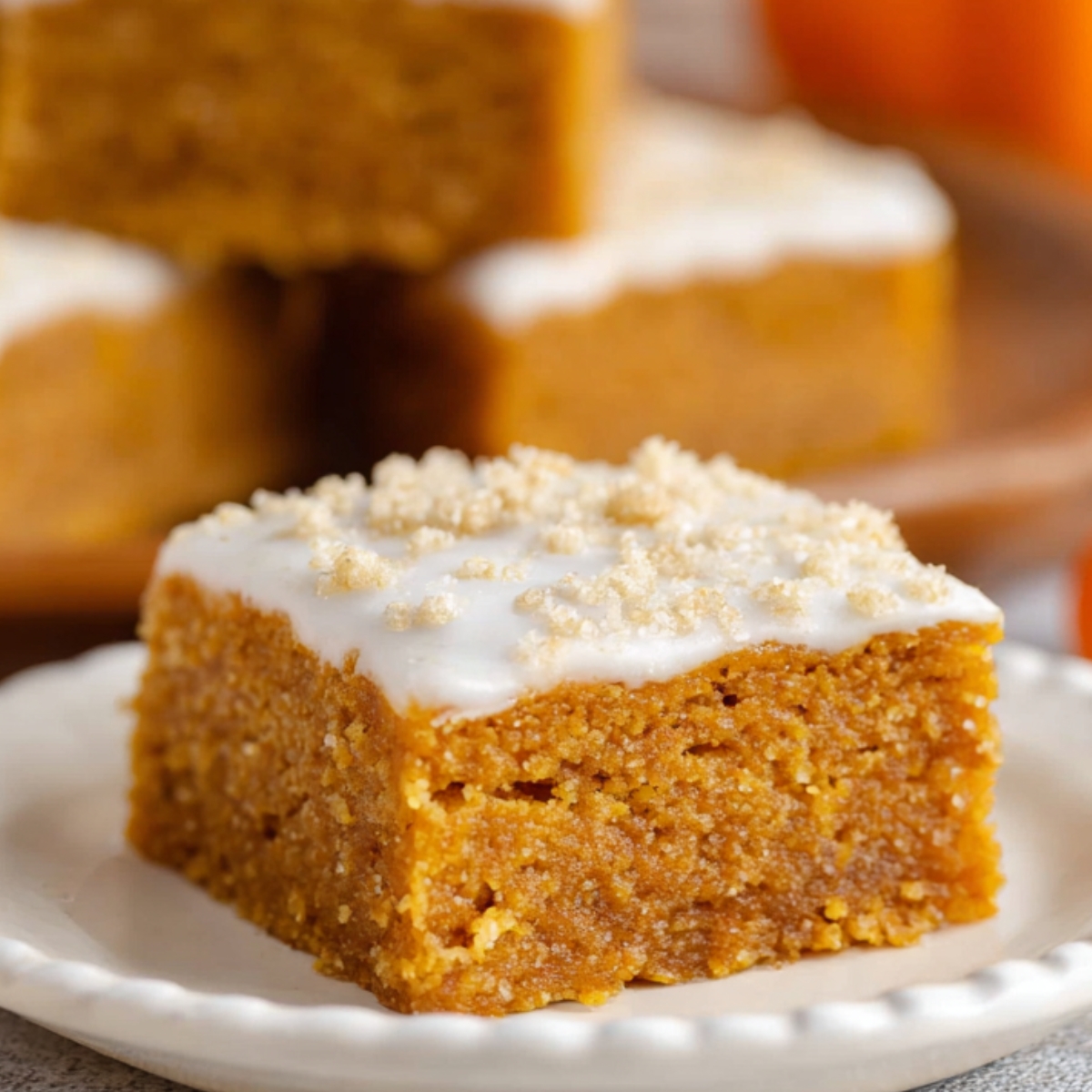 Moist homemade pumpkin bars topped with white frosting and sugar sprinkles, served on a scalloped ceramic plate with other bars in the background.