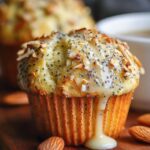 Homemade Almond Poppy Seed Muffins with sliced almonds and glaze, in a paper liner on a wooden board with whole almonds and a coffee mug in the background.