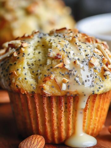 Homemade Almond Poppy Seed Muffins with sliced almonds and glaze, in a paper liner on a wooden board with whole almonds and a coffee mug in the background.