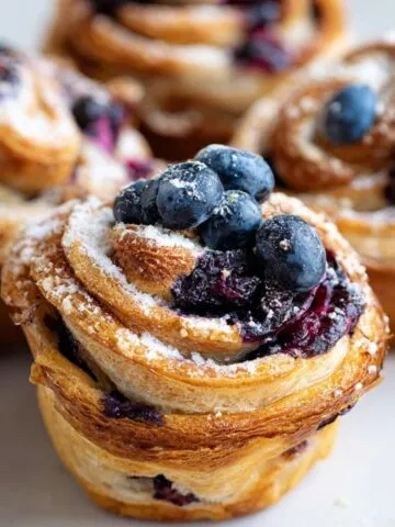 Homemade blueberry cruffins with golden flaky layers, fresh blueberries on top, and a dusting of powdered sugar, shot in natural daylight on a light surface.