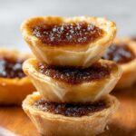 Homemade Butter Tarts with golden flaky pastry and glossy brown sugar filling, on a wooden surface with blurred tarts in the background.
