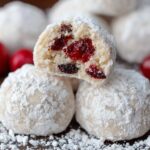 Homemade cherry snowball cookies coated in powdered sugar, one broken open to reveal red cherry pieces, with scattered sugar and whole cherries nearby.