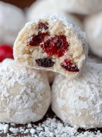 Homemade cherry snowball cookies coated in powdered sugar, one broken open to reveal red cherry pieces, with scattered sugar and whole cherries nearby.
