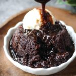 Homemade chocolate pudding cake in a white ramekin, topped with a scoop of vanilla ice cream, as thick chocolate sauce is being poured over the top, pooling into the rich, fudgy cake below.