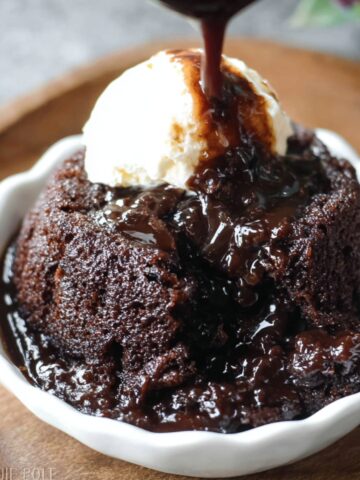 Homemade chocolate pudding cake in a white ramekin, topped with a scoop of vanilla ice cream, as thick chocolate sauce is being poured over the top, pooling into the rich, fudgy cake below.