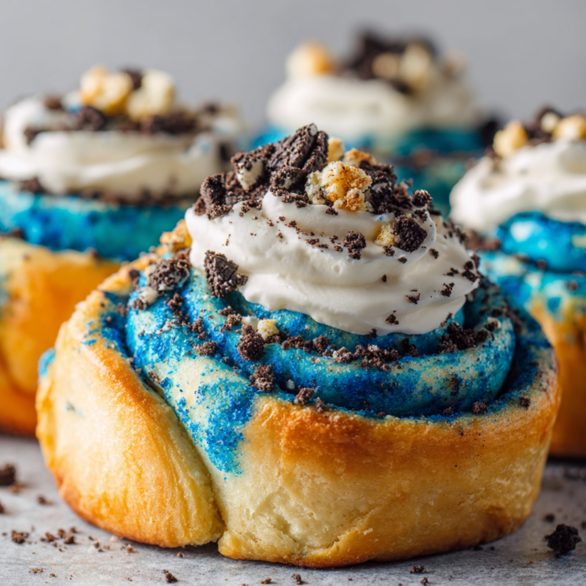 Homemade Cookie Monster Cinnamon Rolls with golden edges, bright blue swirls, a dollop of white whipped cream, and crushed chocolate and vanilla cookies on top. Set on a gray surface with more rolls in the background.