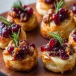 Homemade Cranberry Brie Bites with golden puff pastry, melted brie oozing out, cranberry topping, and rosemary sprigs served on a wooden board.