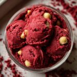 Homemade edible red velvet cookie dough scooped into a glass bowl, with a rich red color, creamy texture, and white chocolate chips throughout. Crumbs are scattered around on a light surface, giving it a fresh, homemade look.
